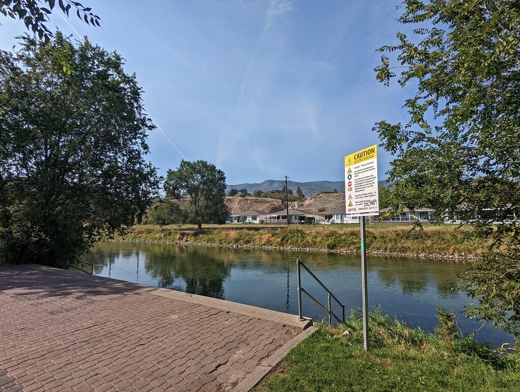 Paved area next to Penticton Channel, with Caution signage and calm water behind. There are houses visible on the other side of the channel