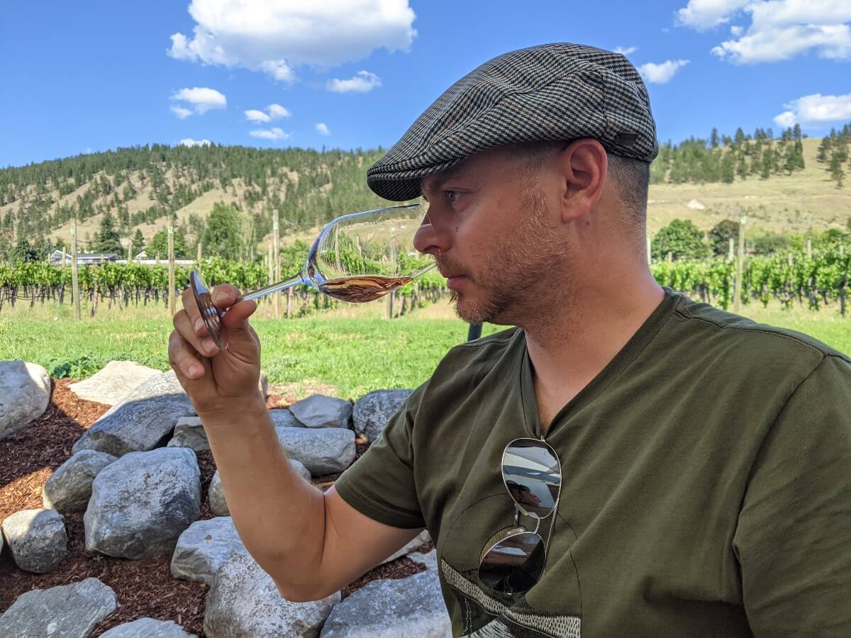 Side view of a man wearing a flat cap hat and sniffing inside a wine glass filled with rose wine, in front of a vineyard