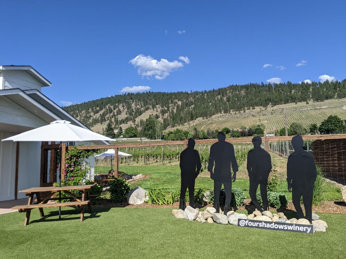 Side view of Four Shadows tasting room, with picnic table (with umbrella) next to life size silhouettes of four young men, in front of vineyards