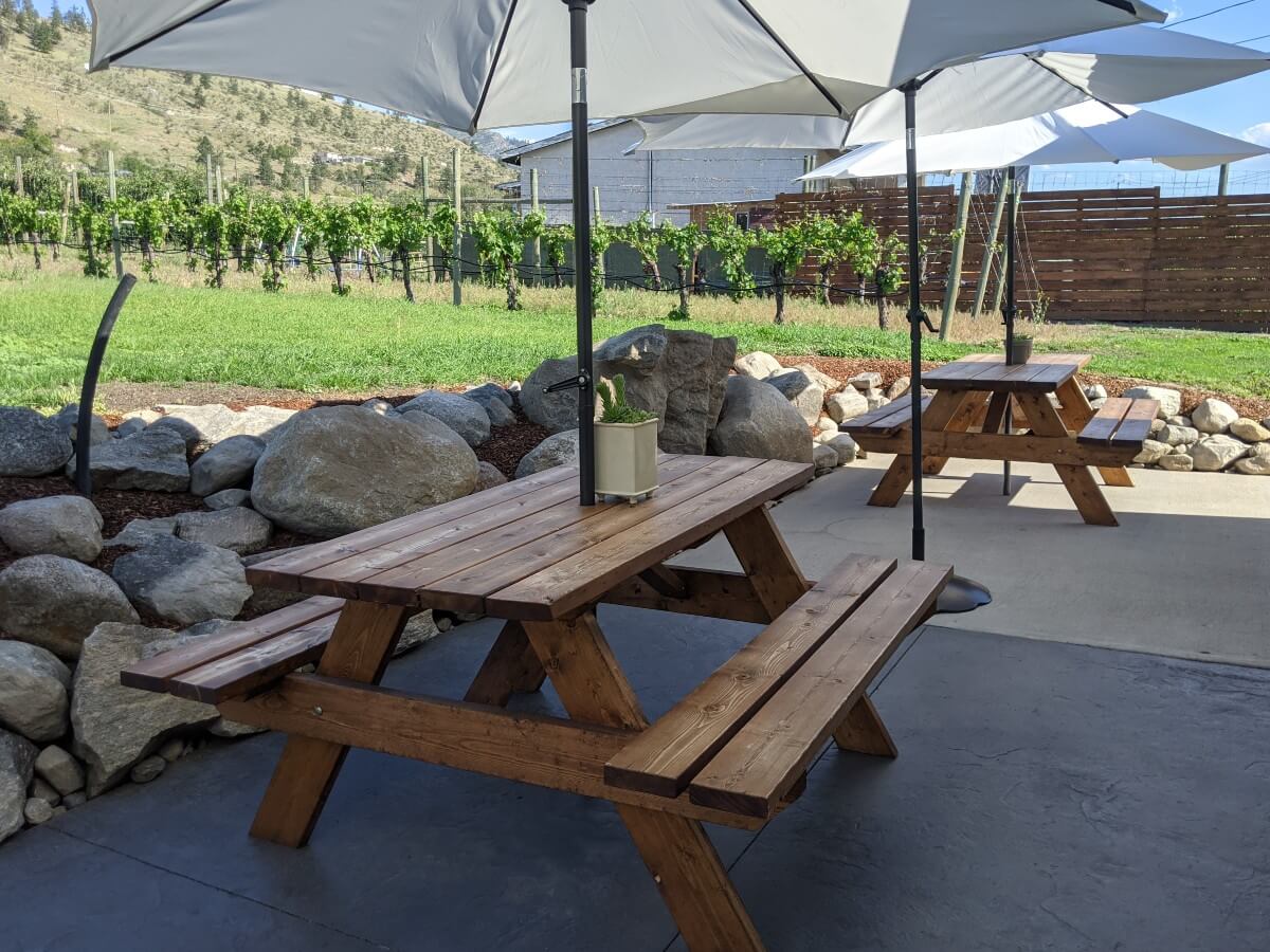 Side view of two wooden picnic tables with open umbrellas on paved patio. The patio is backed by lawn and vineyards