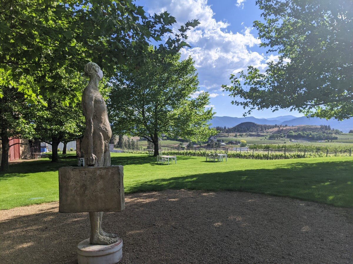 A statue of a man holding a briefcase stands in front of a lawn area with picnic tables and views of vineyards and hills behind