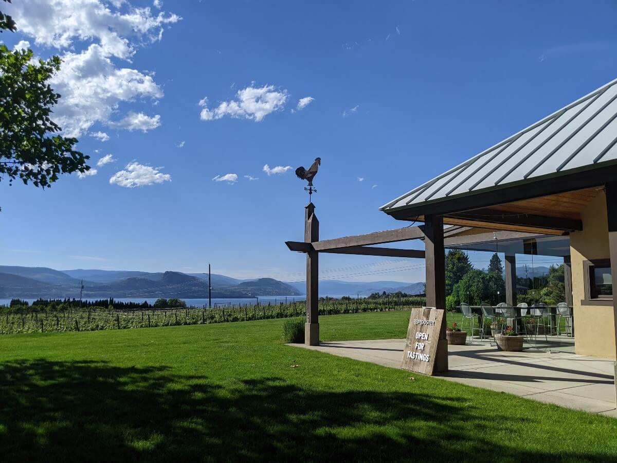 Side view of Red Rooster wine shop and tasting patio, with covered patio area, manicured lawn and lake and vineyard views in the background