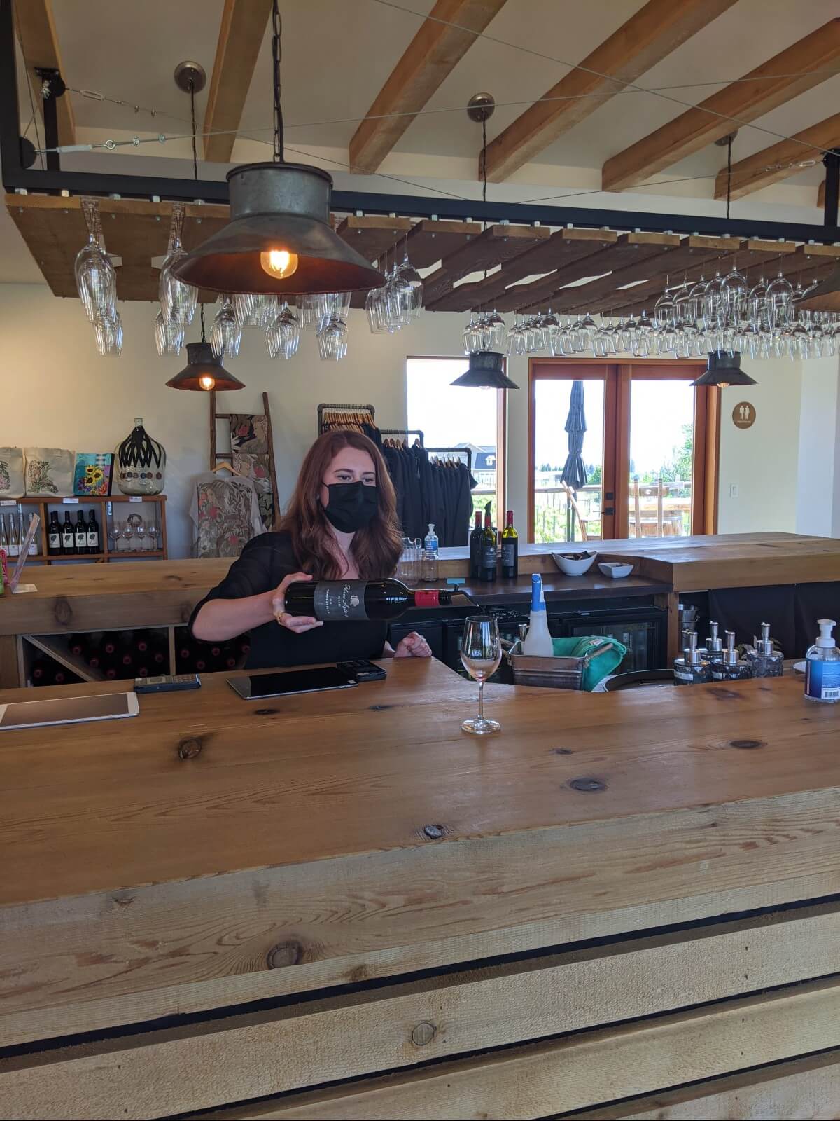 A wine server is pouring red wine into a glass at the wooden tasting bar at Three Sisters Winery, where wine glasses hang from the ceiling