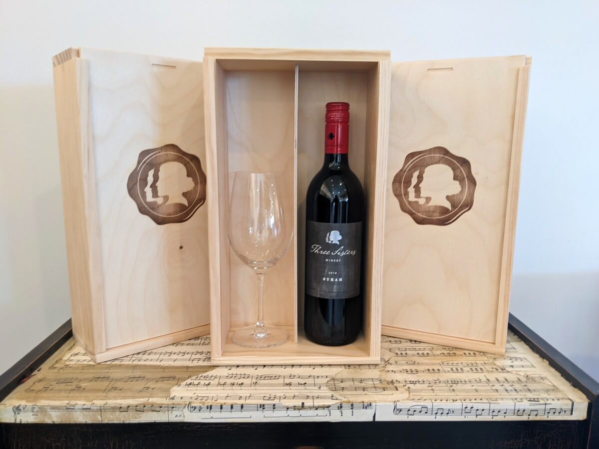 Front view of wooden box with Three Sisters wine bottle with glass, on top of a cabinet