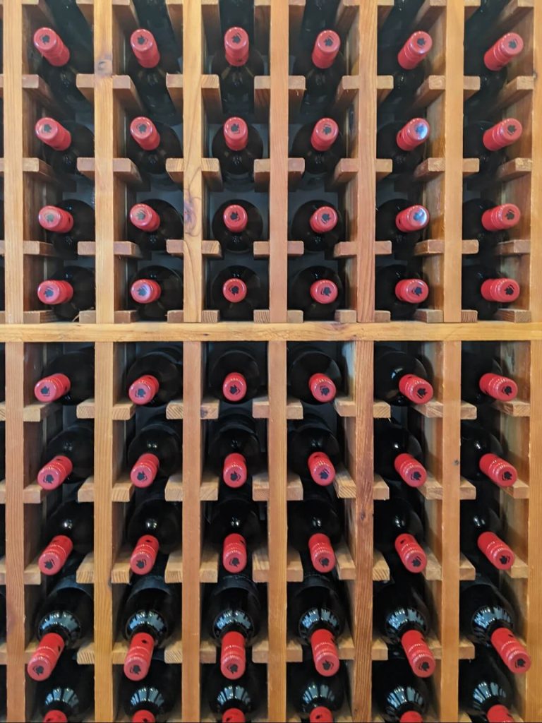 Large wooden wine rack full with bottles. All the bottles are dark in color and all have a red caps with a silhouette of three women stamped in black on the top or the each red caps. 
