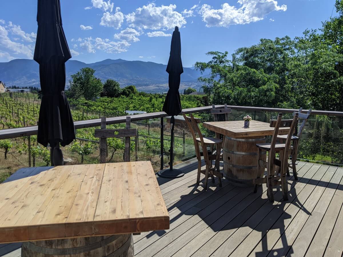 Side view of two wooden tables (with wine barrels as support) on wooden patio in front of vineyards at Three Sisters Winery