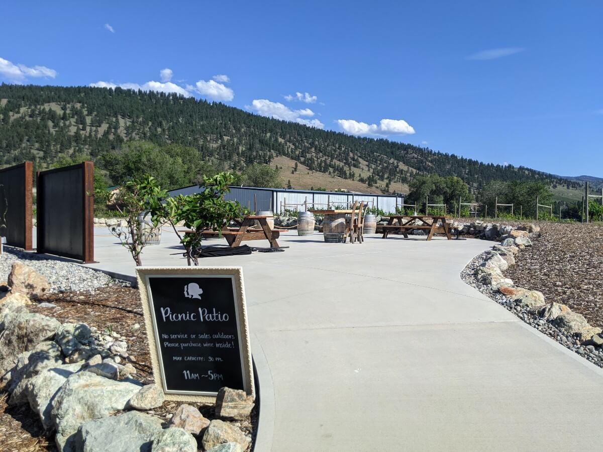 Looking up towards paved area with picnic tables and wine barrels, lined by rocks, with forested mountain in background