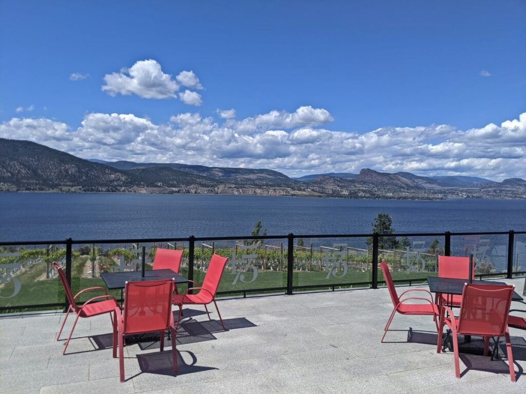 Red chairs and tables on patio at Bench 1775 with Okanagan Lake and vineyards visible in background, backdropped by forested mountains
