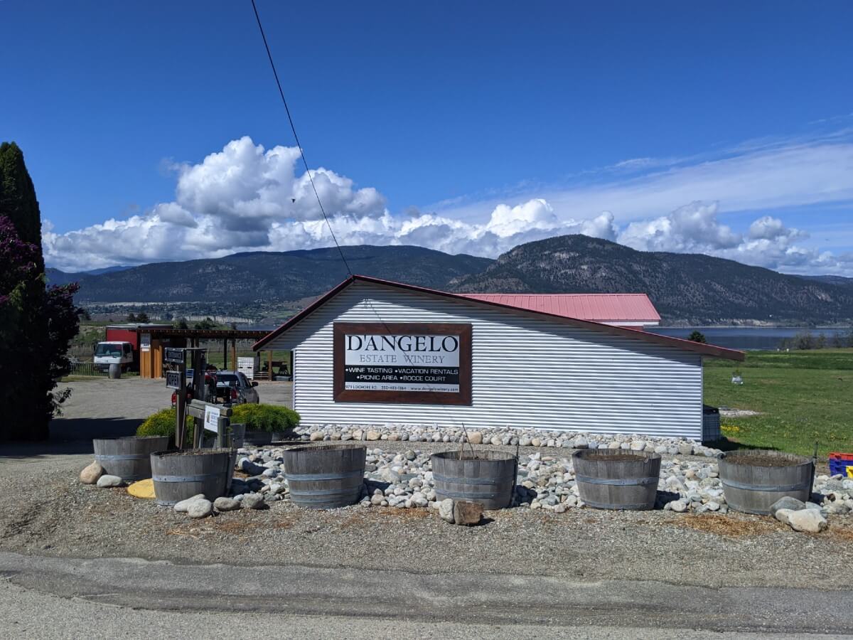 Road view of D'Angelo Winery, with white building featuring D'Angelo logo with adjacent parking lot, surrounded by grass, with peek through view of Okanagan Lake in background
