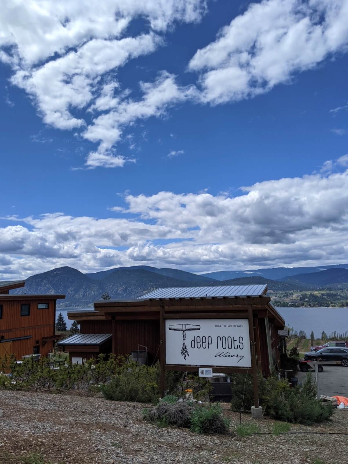 Deep Roots Winery building, with large white sign featuring logo, in front of small wooden building, with Okanagan Lake visible behind