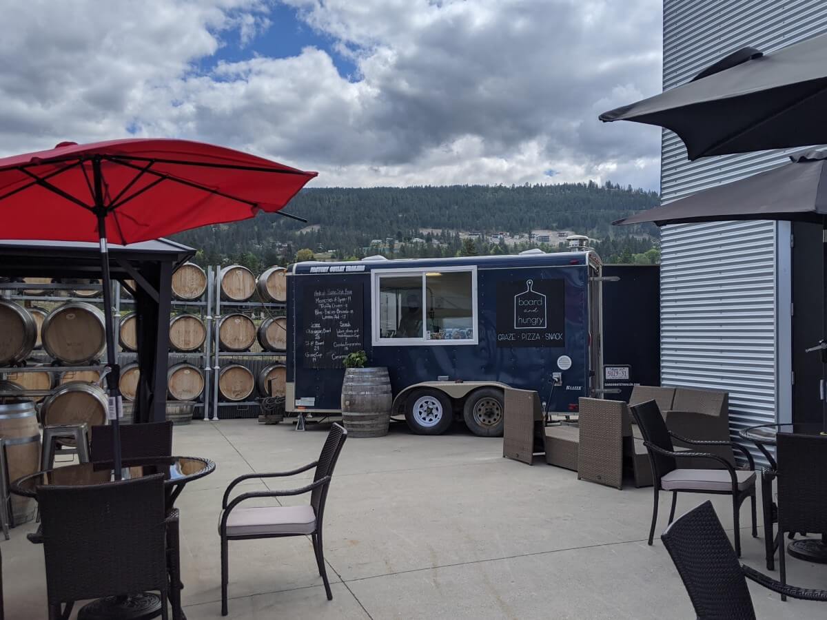 View of Therapy Vineyards patio with multiple kinds of seating, with small blue food truck in background, with Board and Hungry sign