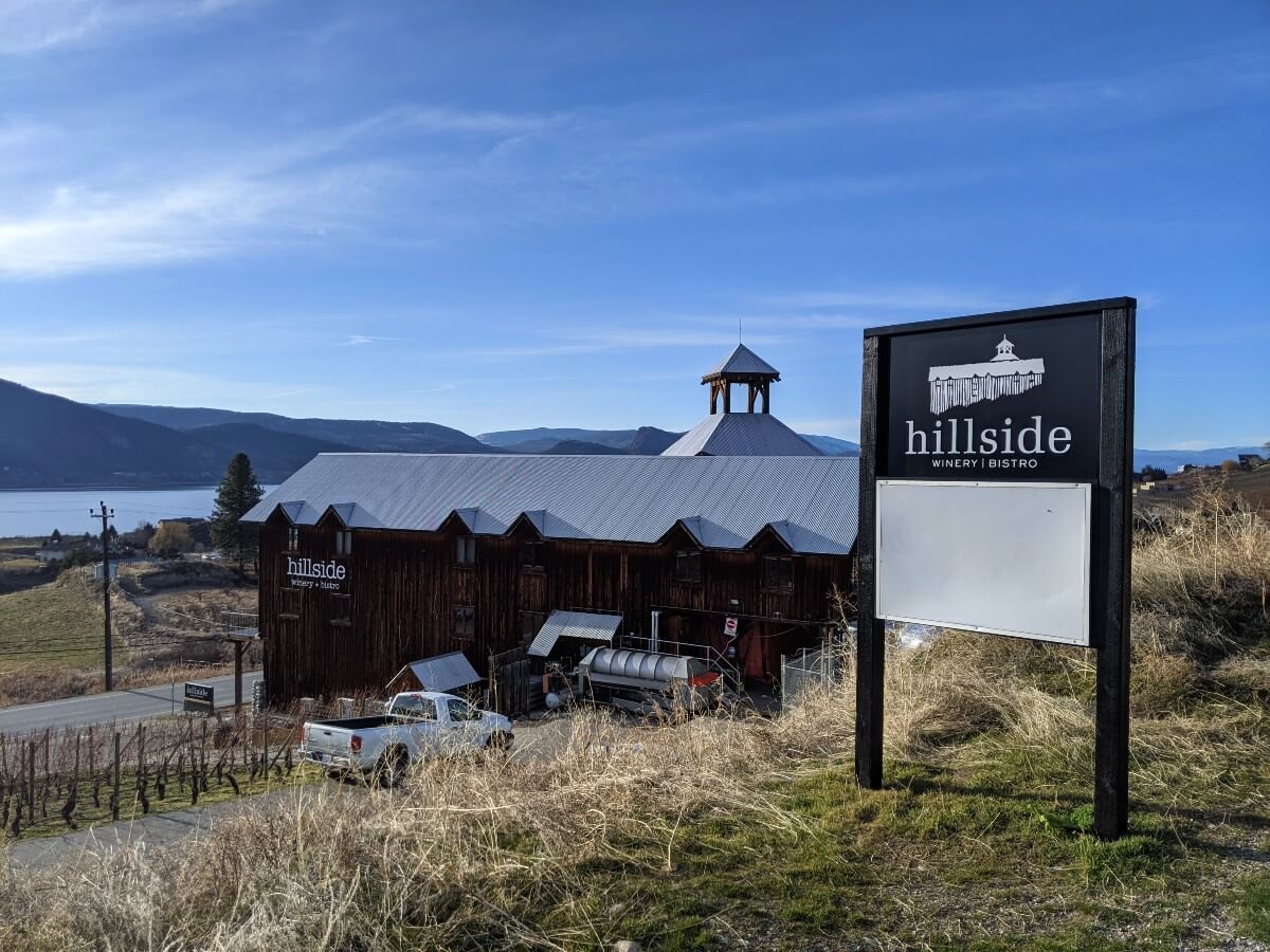 View looking down from hiking trail towards Hillside Winery, a tall brown building next to Naramata Road. There is signage to the right