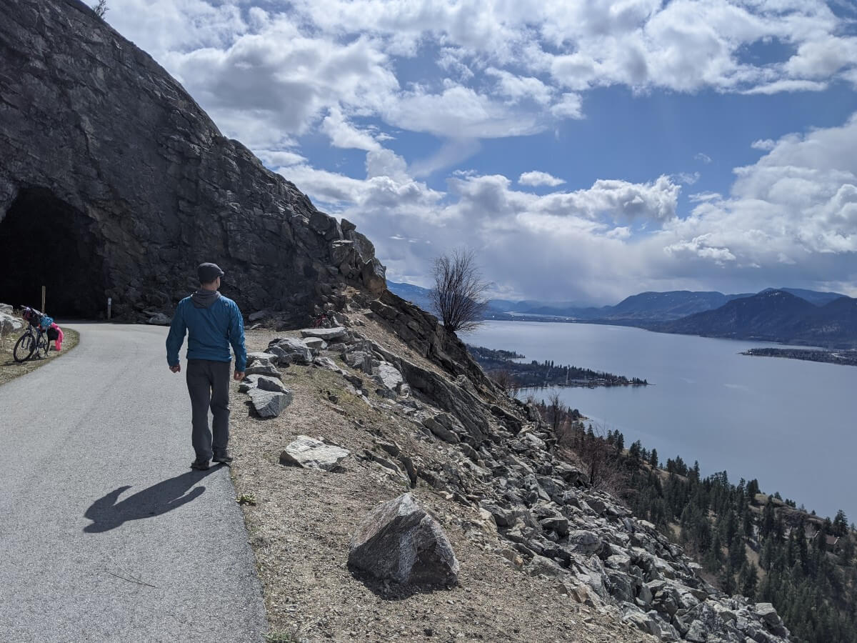 Back view of man walking along paved hiking path leading towards tunnel, with lake visible below