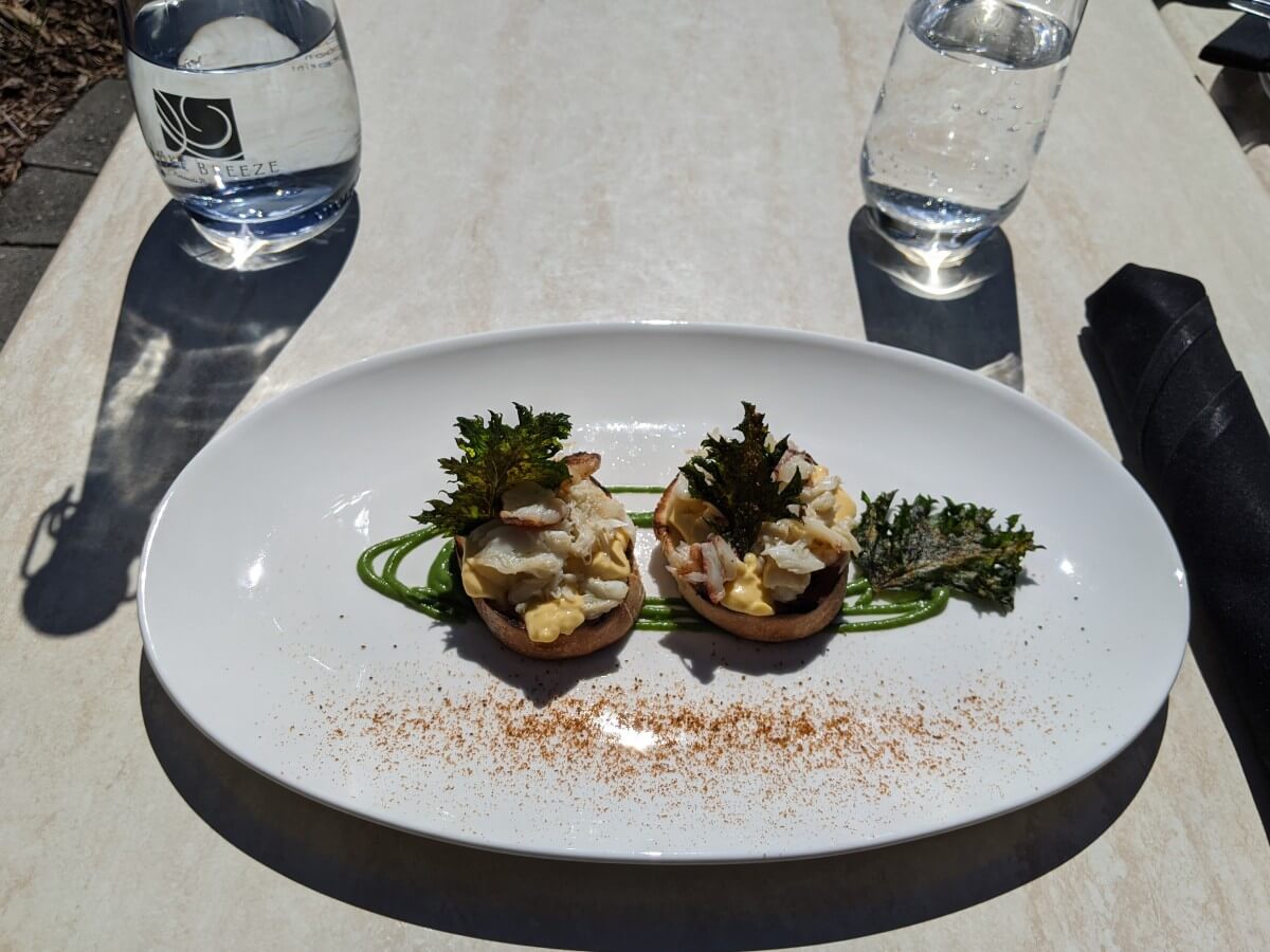 Looking down on white plate on table, with two round toasts topped with crab, custard and crispy kale. There is a green sauce piped underneath the bread. There are two glasses set behind the plate.