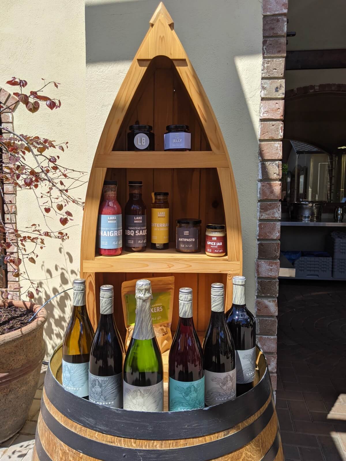Six full bottles of Lake Breeze wine sit on top of a wine barrel, with shelving behind with jarred conserves and sauces
