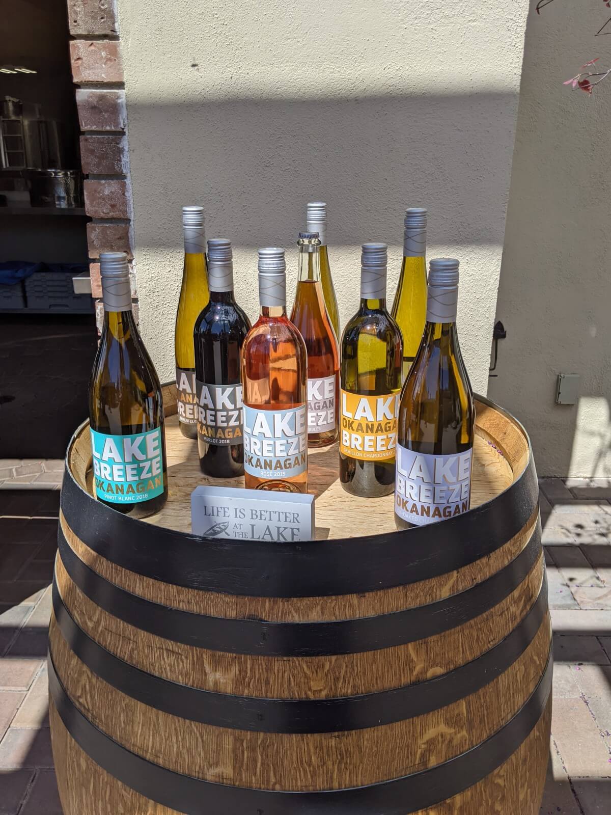 Nine Lake Breeze wine bottles stand on top of a wine barrel (multiple different colours and shapes), with a sign 'Life is Better at the Lake' in the foreground