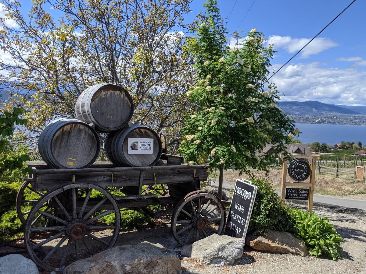 View of Mocojo Winery entrance with signage and wooden cart with wine barrels. Okanagan Lake is visible in the background