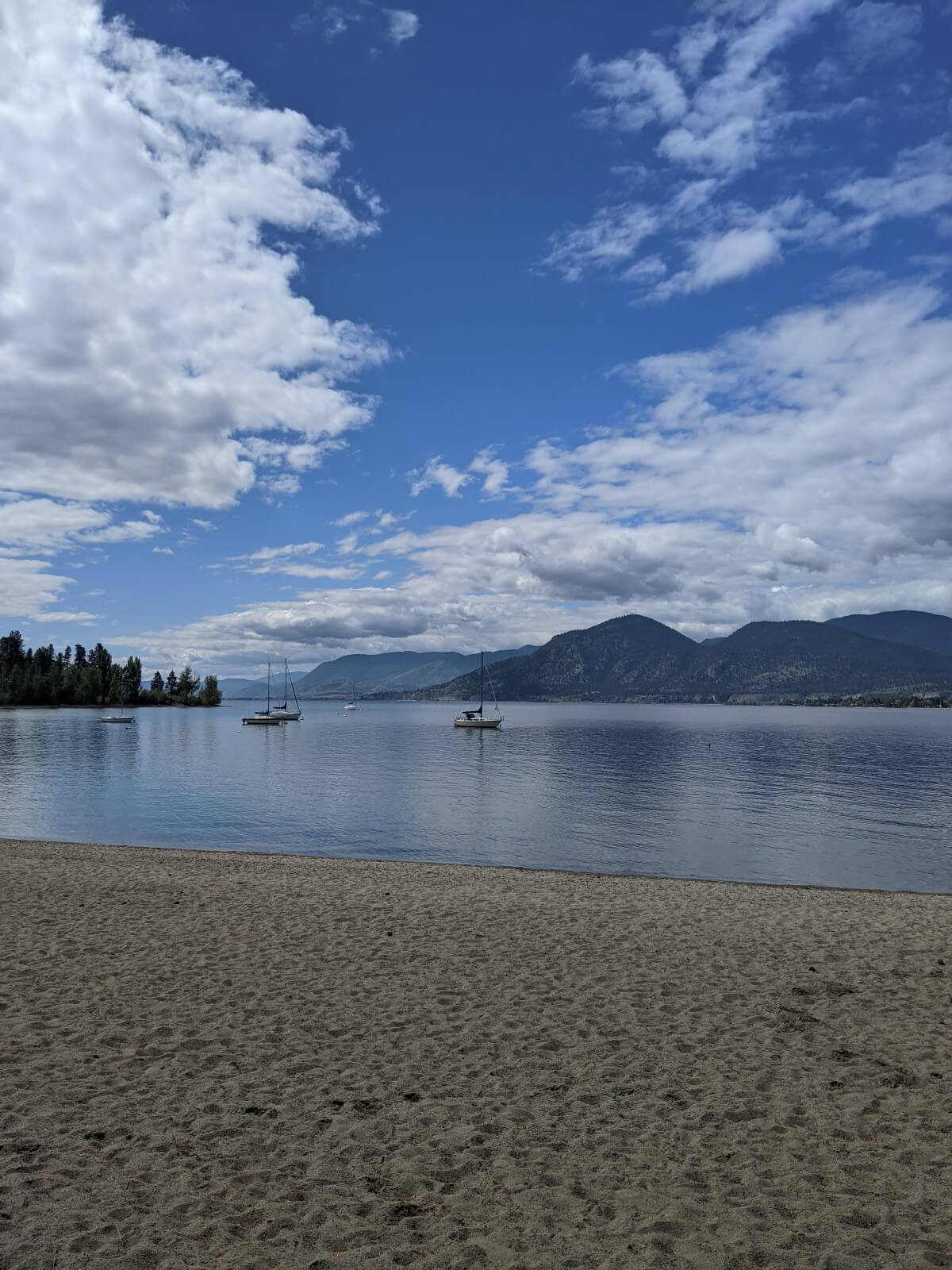 View of sandy beach next to calm lake backdropped by rounded mountains, with four boats on the water