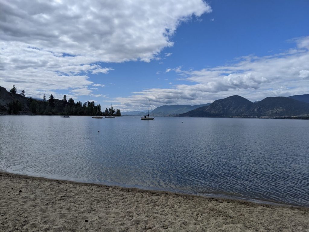 View from Manitou Beach looking out onto calm Okanagan Lake, where several boats are moored. Forested mountains are visible on the other side of the lake