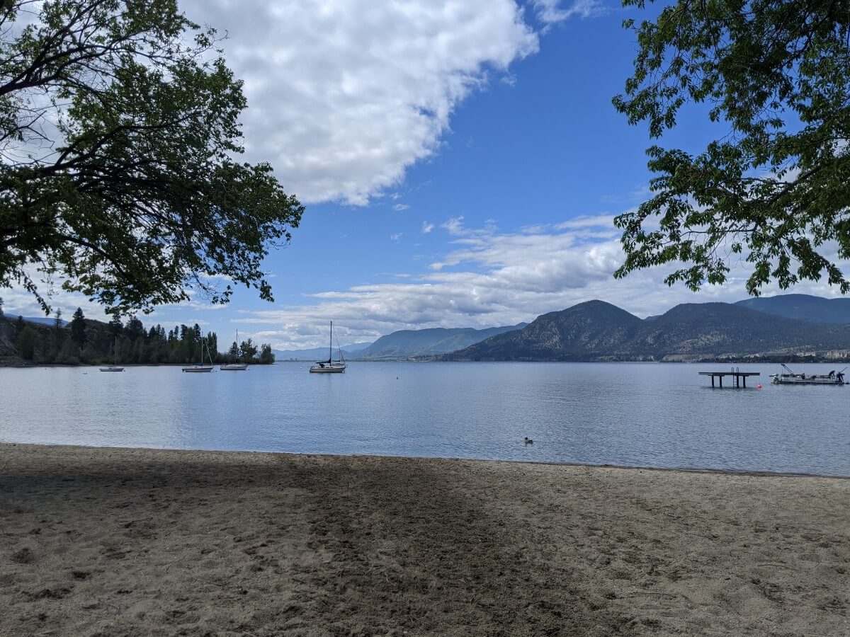 View of Manitou Park beach with golden sand in front of calm lake. A duck swims past near the shore, while a handful of boats sit just offshore. There are forested hills on the other side of the lake