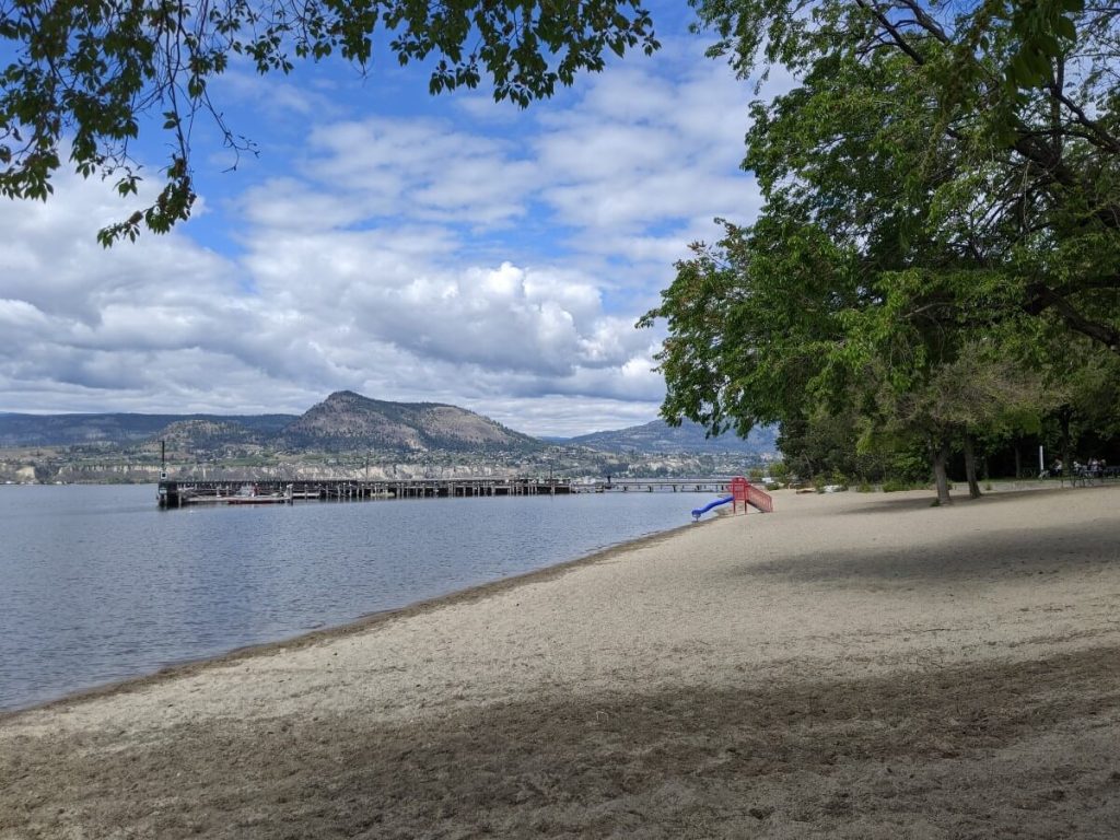 Side view of Manitou Park beach with golden sand in front of calm lake, framed by trees. There is a dock visible in the background. A mountain rises above the lake. 