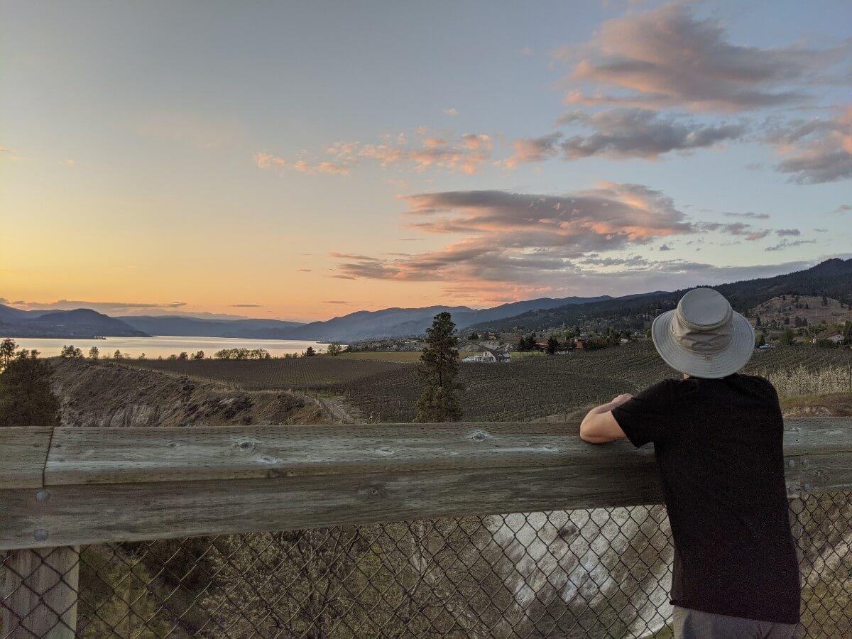Back view of man leaning over wooden fence on the McCulloch Trestle looking out to pretty sunset over vineyards and lake view