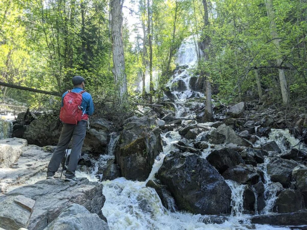 Back view of man with a red backpack and blue jacket standing on a rock looking up at tall waterfall flowing down in between  large boulders. 