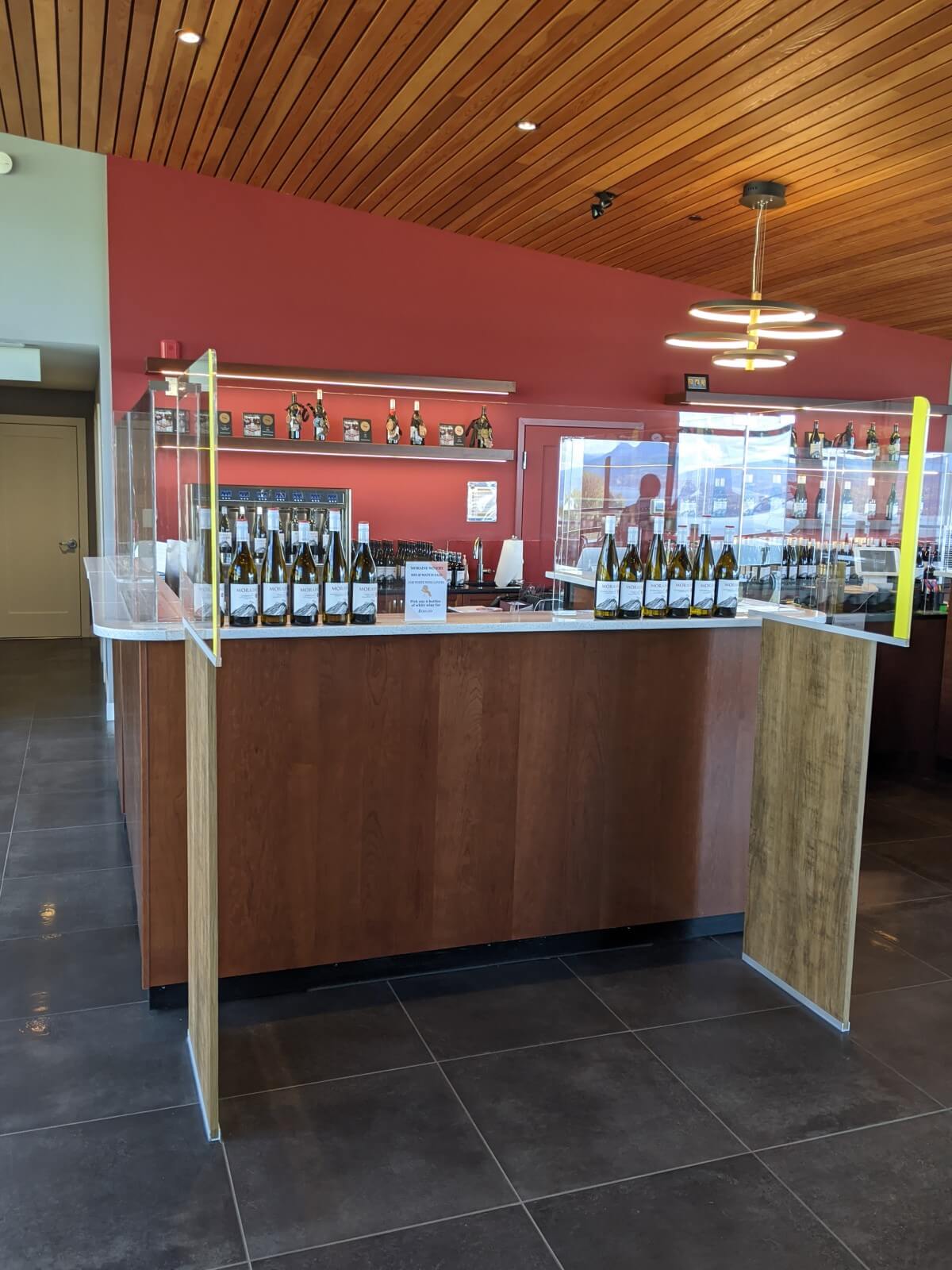 Front view of inside tasting bar at Moraine Winery, with wooden ceiling, red painted wall and wooden counter. The sides of the counter have plexiglass barriers. There are twelve bottles of wine lined up on the counter