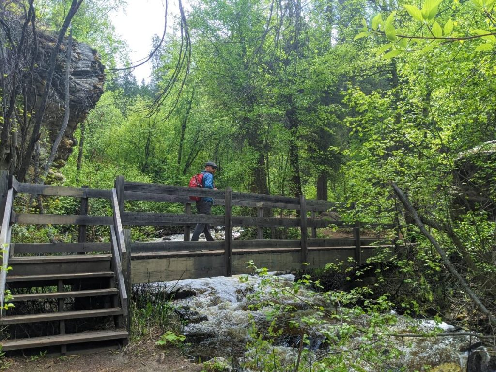 Side view of man with red backpack crossing wooden bridge on Naramata Creek Falls. The rushing water below the bridge is visible. Forest surrounds the bridge