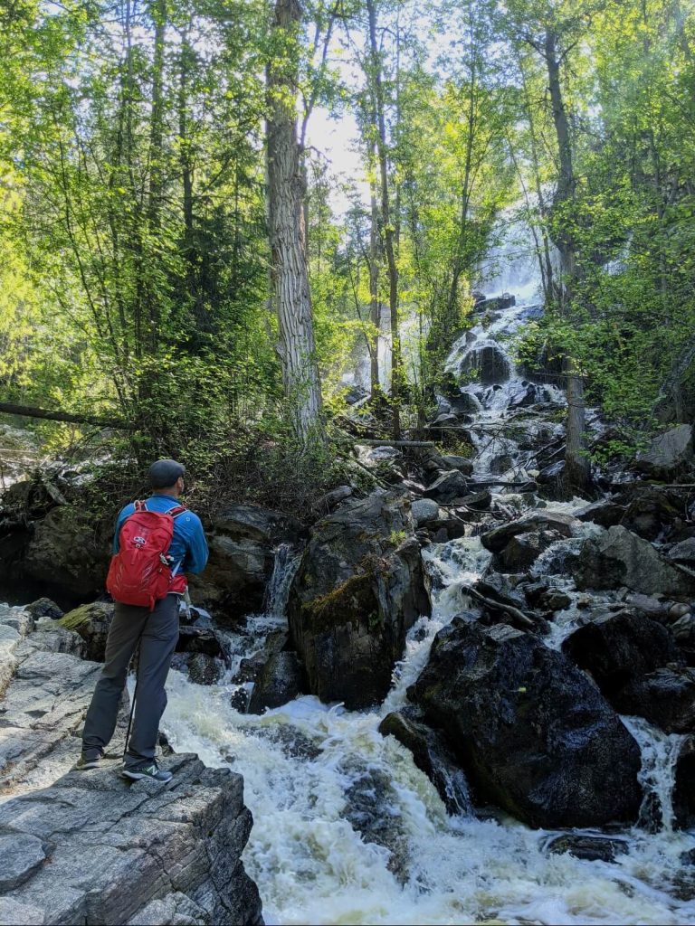 Back view of man with red backpack looking up at Naramata Creek cascade, which is a multi-layered waterfall surrounded by forest