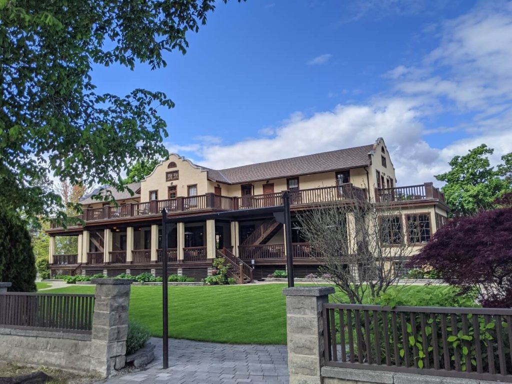 Front view of the Naramata Inn, a two floored cream coloured building with brown fenced balconies running on the outside. There is a large lawn in front, behind stone gates