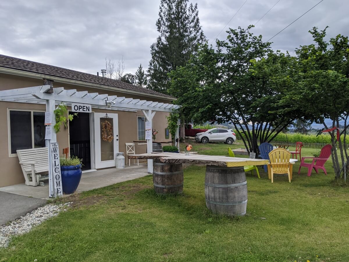 Side view of brown single story tasting room with open sign. Outside there is a rustic wooden tasting bar with colourful Adirondack seating visible to the right