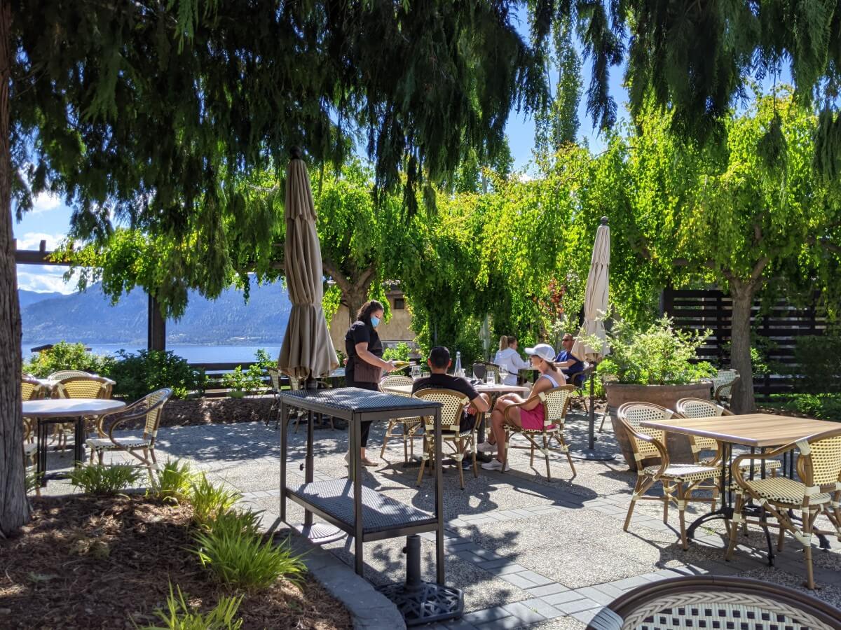 View of restaurant at Lake Breeze, with paved patio with scattered tables, bordered by trees and plants