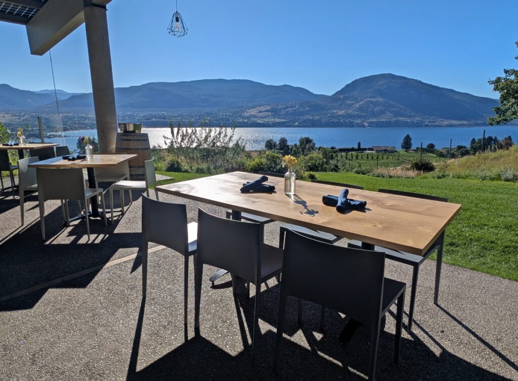 Two tables on the patio at the Poplar Grove restaurant, with views of Okanagan Lake behind, backdropped by forested mountains