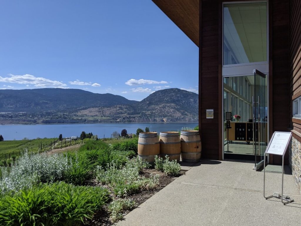 Side view of the entrance to Poplar Grove Winery, with large glass doors leading into an airy, two story building with floor to ceiling windows. Next to the door is three wine barrels and a sign. There are views of Okanagan Lake and mountains to the left.