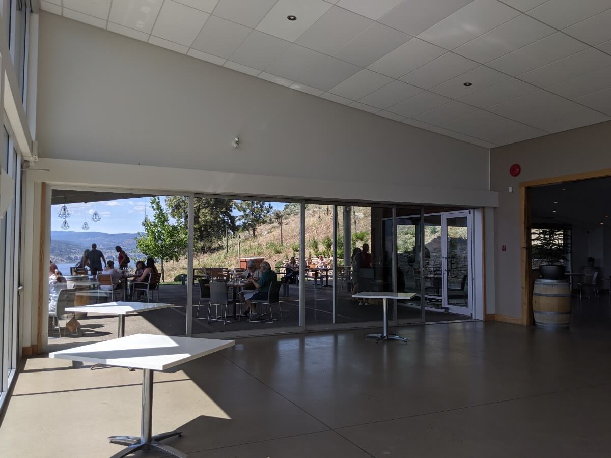 Side view of dining patio at The Restaurant at Poplar Grove, through glass windows. There is an outdoor patio with spaced seating, with views of Okanagan Lake visible in the background