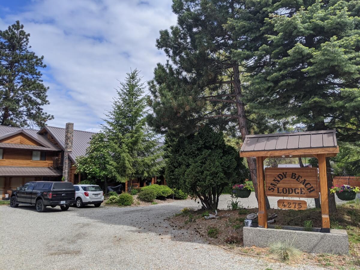 Wooden Sandy Beach Lodge sign on right, with two floor lodge building visible on the left, with two vehicles parked in front 