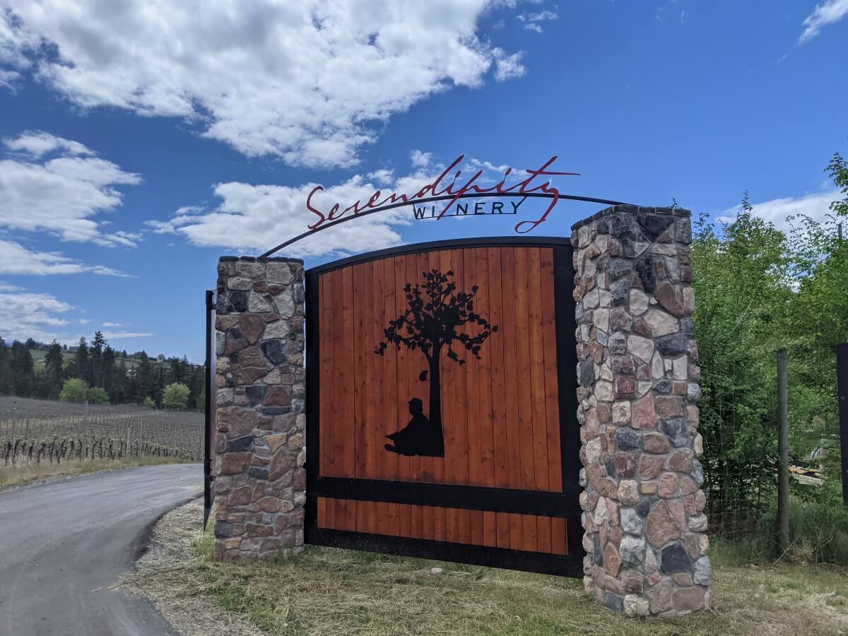 Roadside view of the gate at Serendipity Winery, a large wooden sign with silhouette of man sitting underneath an apple tree, with the winery name above