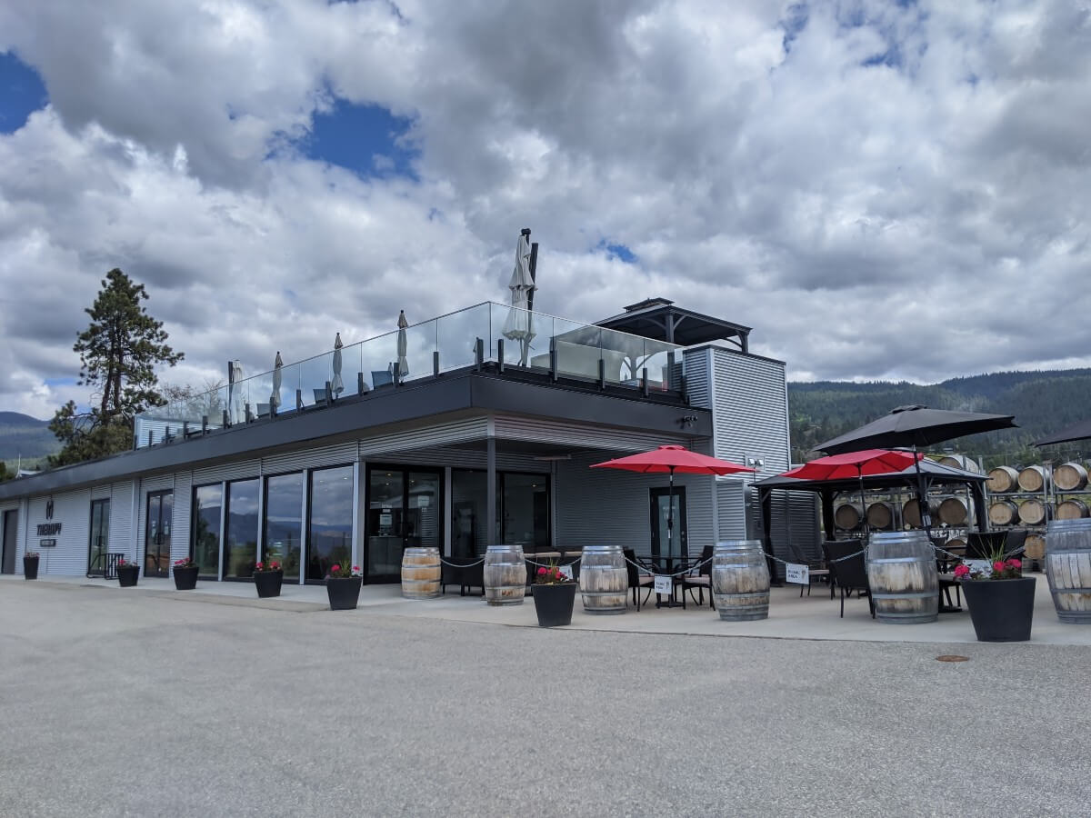 Side view of two story Therapy vineyards tasting room featuring floor to ceiling windows, with red umbrellas and seating outside ground floor and seating on roof top patio
