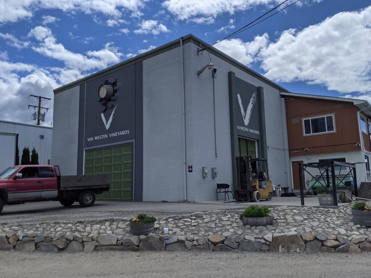 Van Westen Vineyards building exterior with gray warehouse featuring large doors and forklift parked outside, red truck on left. The winery's logo is on both sides of the building