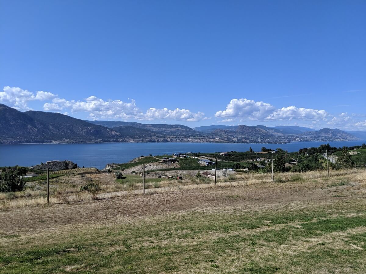 Looking out over a grassy area down to vineyards and houses, in front of Okanagan Lake, which is backdropped by mountains