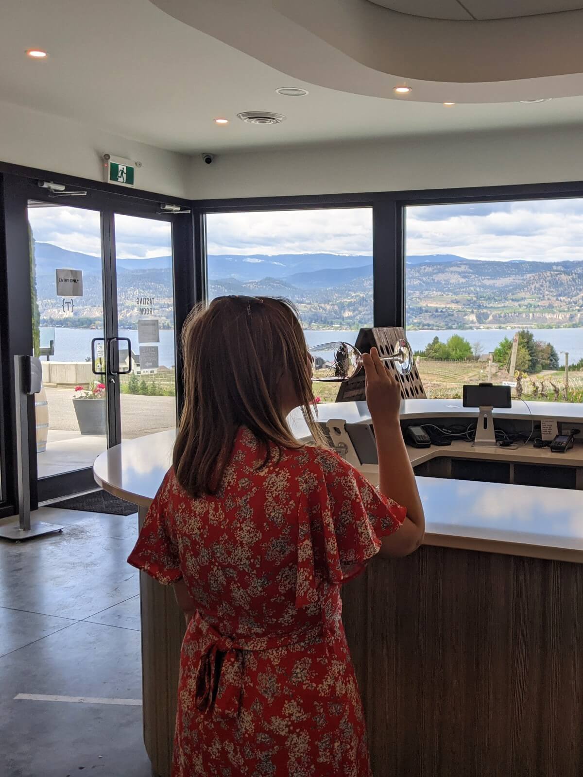 Back view of woman in red dress sipping wine from wine glass at tasting bar, with views of Okanagan Lake visible through floor to ceiling windows behind