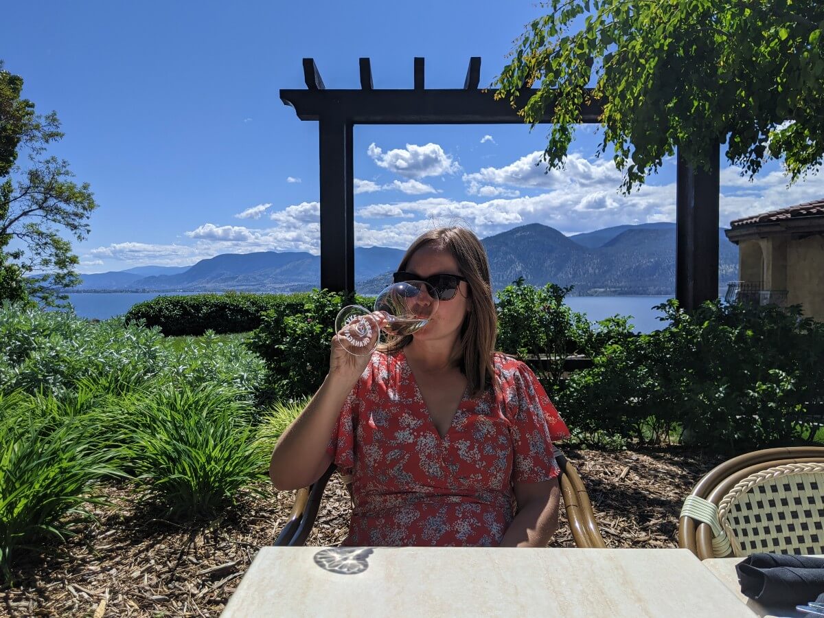 Gemma sits on Lake Breeze patio sipping wine and wearing red dress. Lake and mountain views are visible in the background