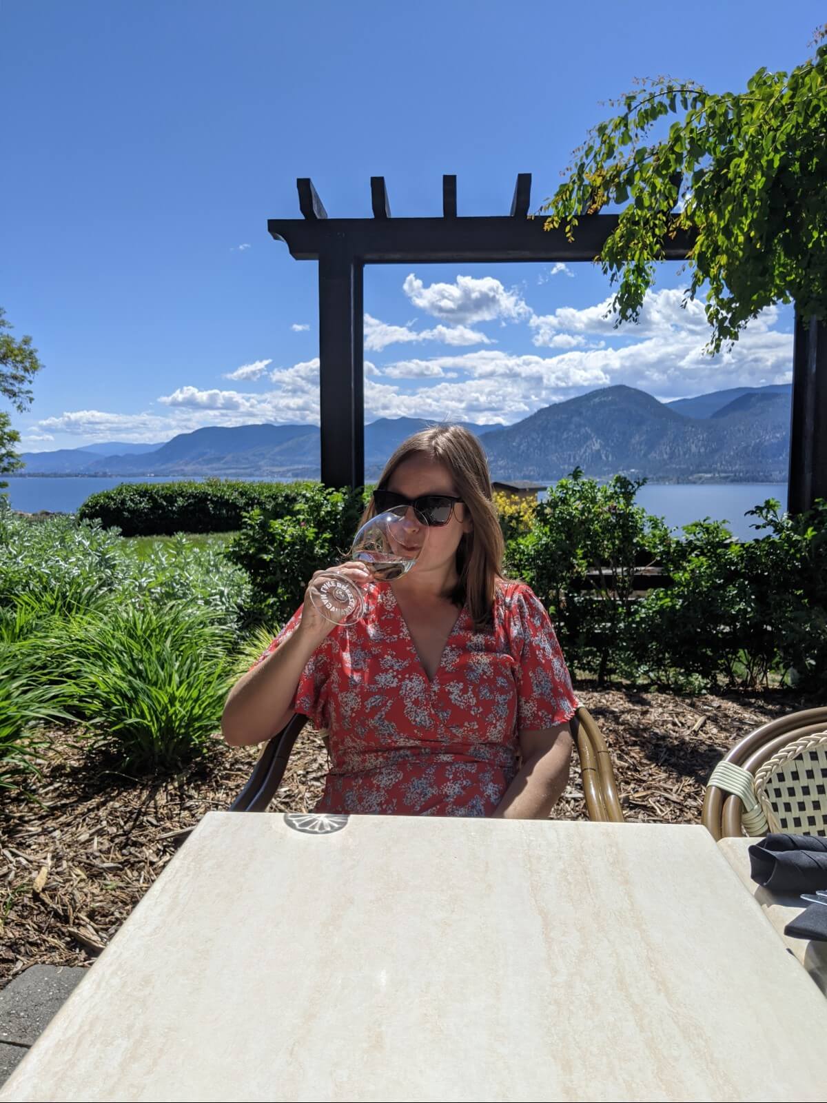Front view of woman in red dress drinking from wine glass at white table, with view of lake and mountains behind