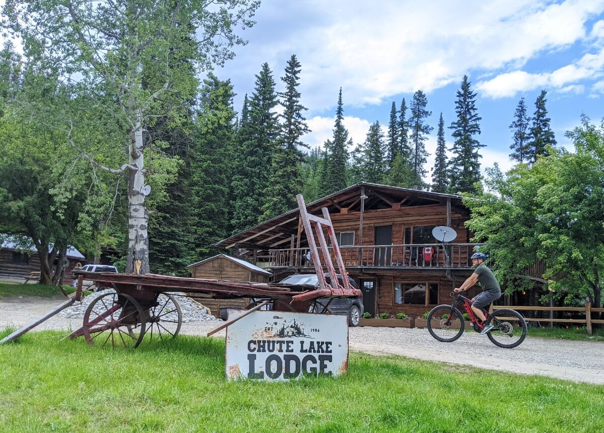A man rides a bicycle in between Chute Lake Lodge signage and Chute Lake Lodge itself, a historic two story wooden building