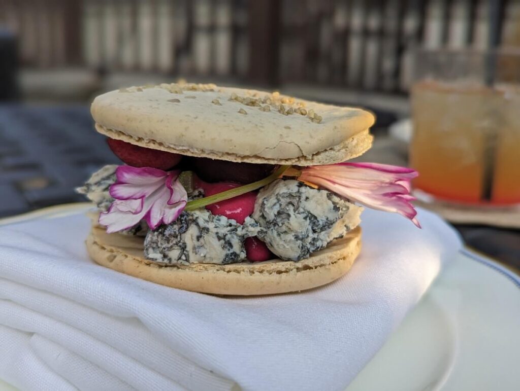 Close up of sunflower macaron, made with locally made Poplar Grove Tiger Blue cheese with orange mocktail glass in background