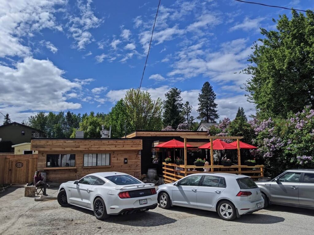 Exterior view of Grape Leaf Cafe with wooden one story building and patio. There are red umbrellas above the tables on the patio. The patio is bordered with trees and many flowers