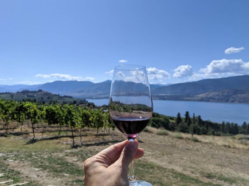 Hand holding glass of red wine in front of camera, in front of views of vineyards and Okanagan Lake