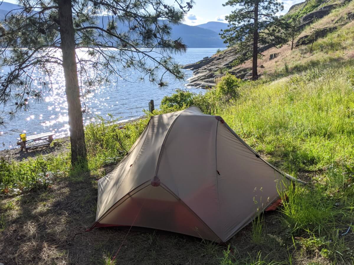 A grey tent is set up on a patch of dirt and grass, overlooking a sparkling lake in Okanagan Mountain Provincial Park near Naramata