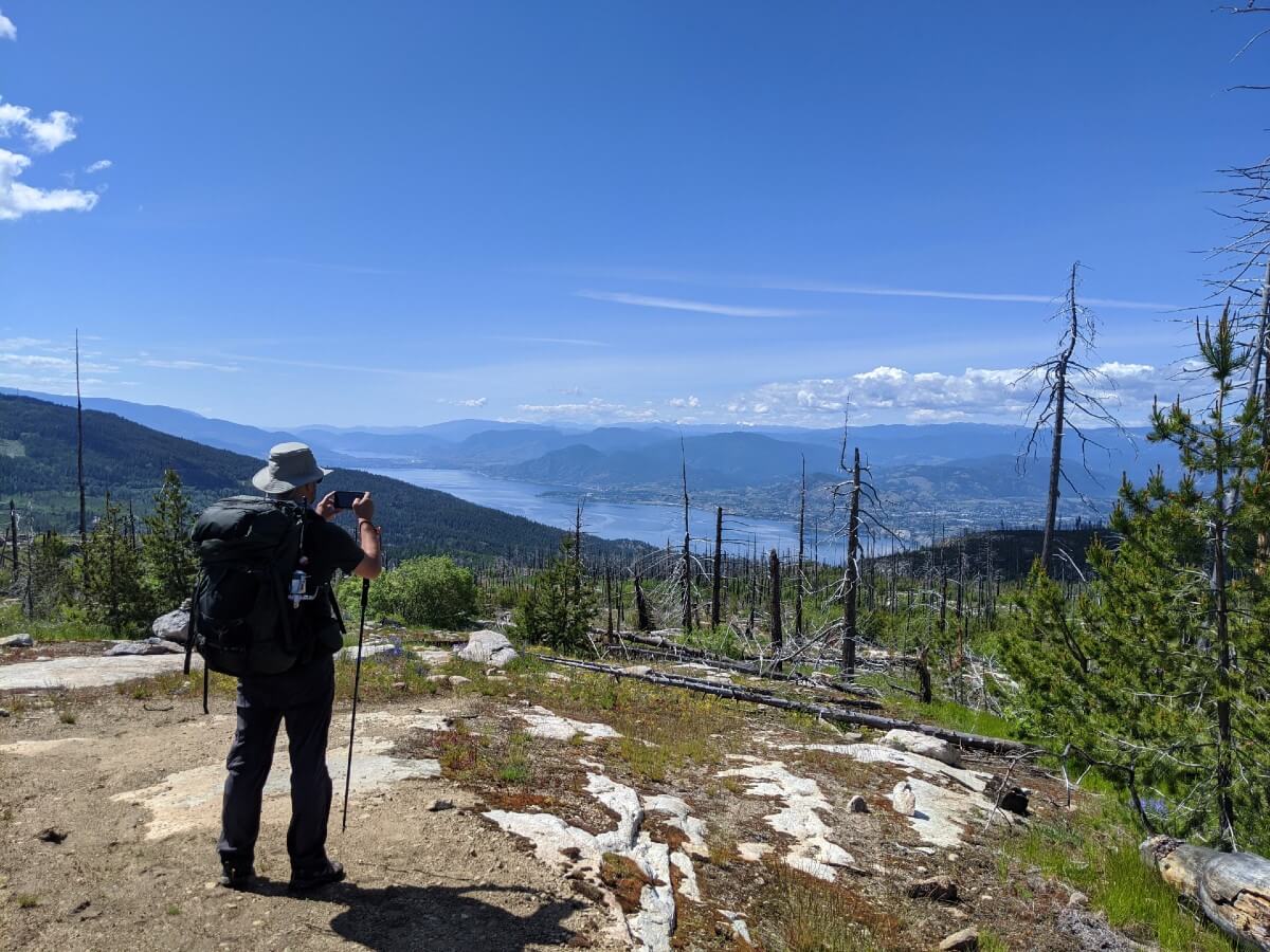 A man with a large backpack stands with his back to the camera, taking a photo of the view of a lake, surrounded by mountains, below